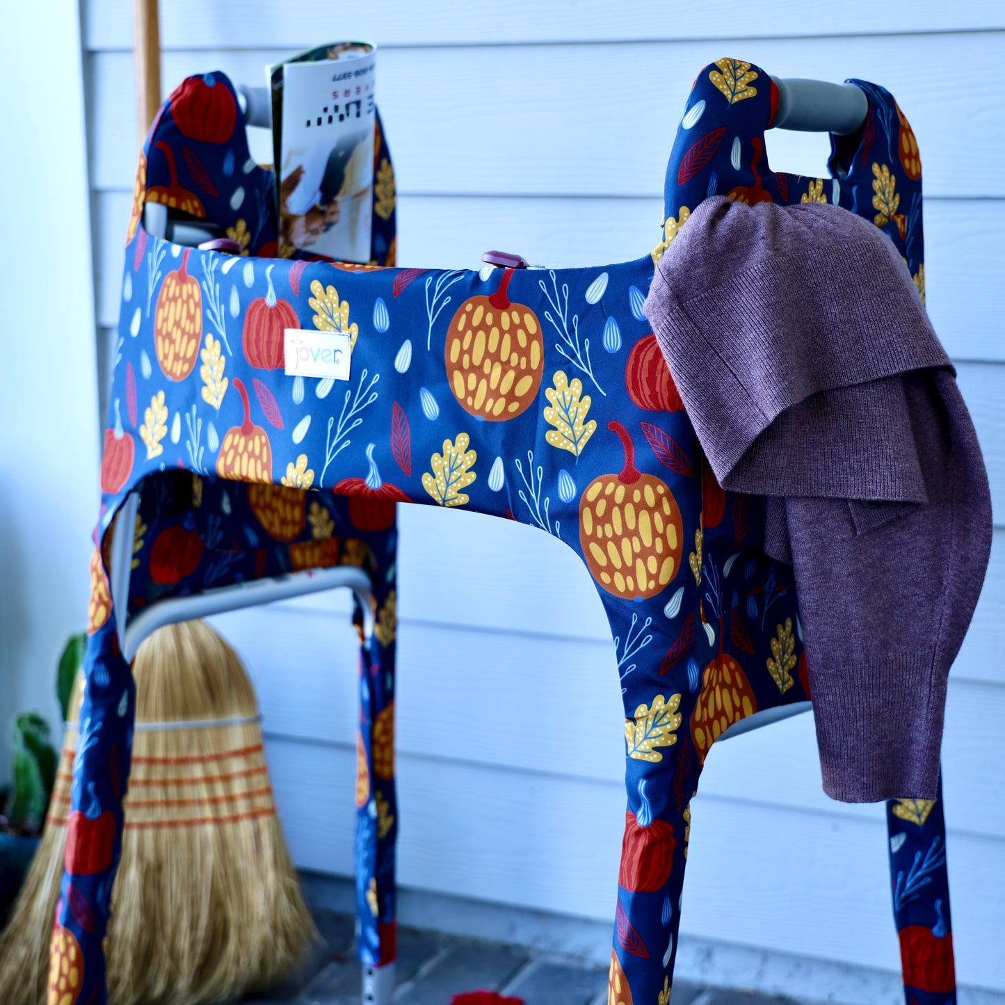 Jover walker cover in Fall Festival (Navy blue with a pumpkin and fall leaf print) on a standard folding walker with a sweater and a magazine in the pockets.