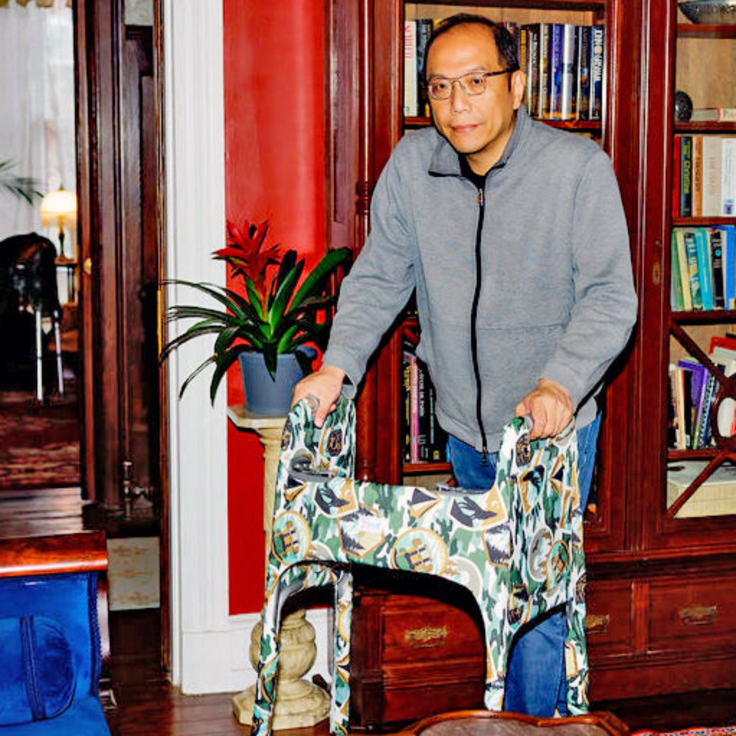 A man standing in front of a bookshelf in a historic home using a metal folding walker outfitted with a Jover Walker Cover in a camping inspired print called the Happy Camper.