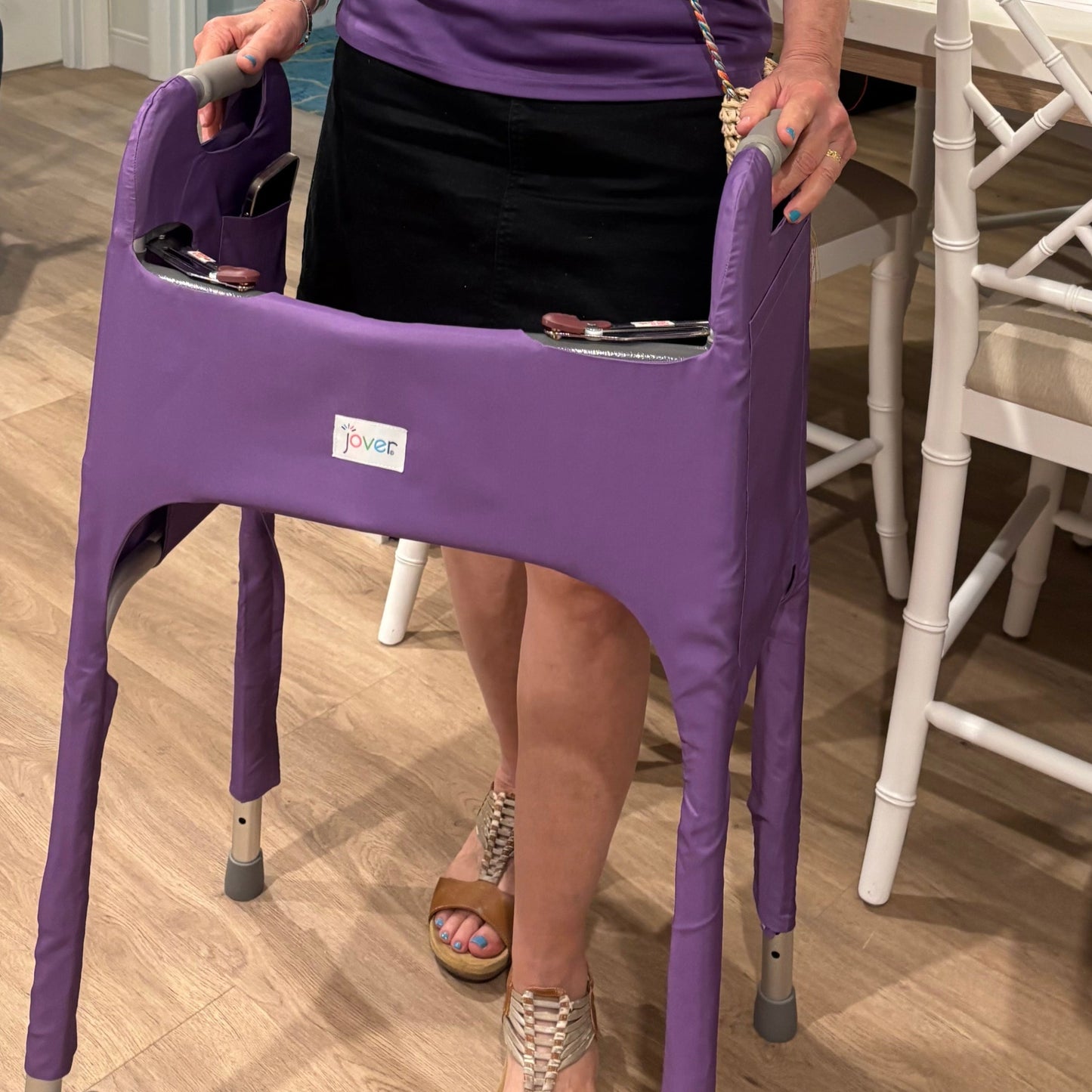Person in a purple shirt in a room with wood flooring, standing behind a standard walker clad in a royal purple Jover walker cover with a phone in the inside pocket.