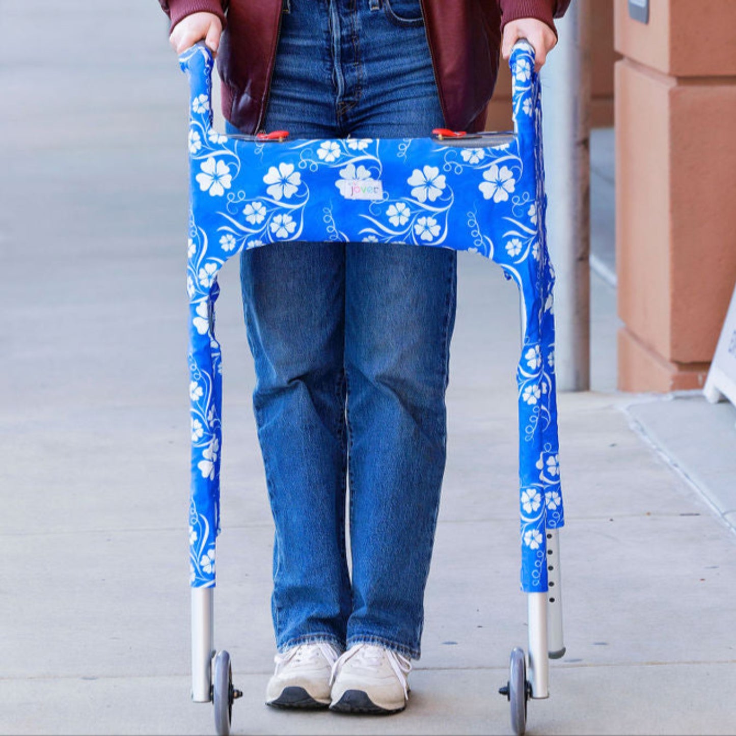 Woman walking behind standard walker clad in a blue and while floral patterned Jover walker cover on a sidewalk with a building in the background