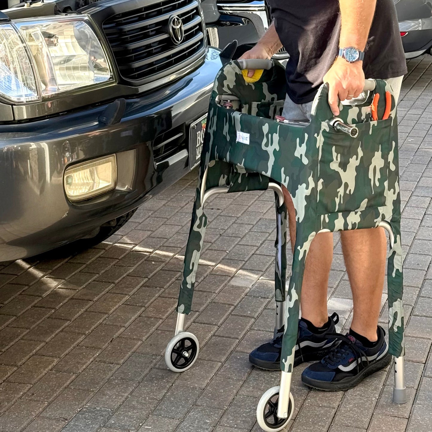 Man working on a car standing behind standard walker clad in Woodland Camo Jover Walker Cover with tools in the outside pocket.