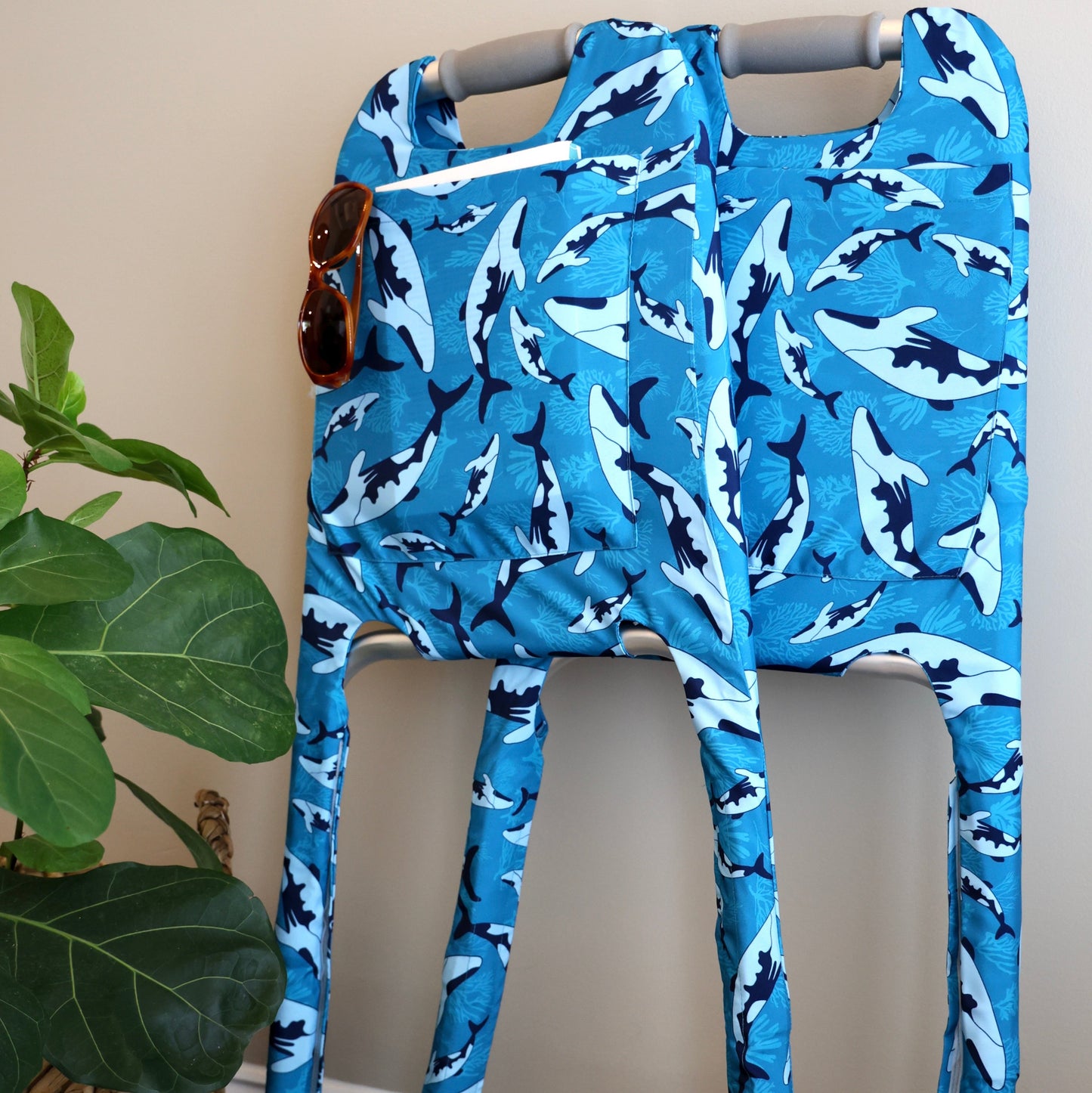 Blue whale patterned Jover walker cover with sunglasses and book in the cover pocket on a folded standard walker leaning against a wall next to a plant