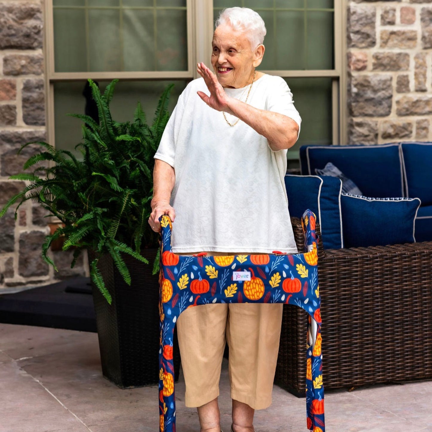Woman on a patio waving and smiling using a walker clad in the Jover Walker Cover in Fall Festival--a dark blue backdrop covered in festive orange and yellow pumpkins and fall leaves. With pockets and style, it's the perfect fall accessory.