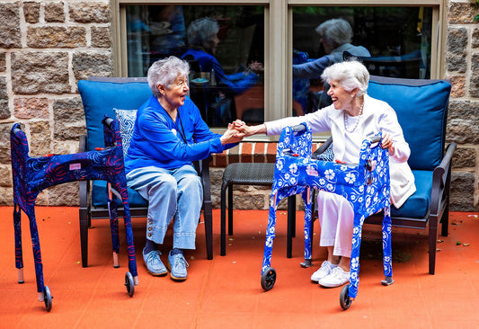 Two women friends seated while socializing happily on a patio. By their sides are walkers clad in Jover Walker Covers. One is a fireworks pattern and the other a blue and white floral.