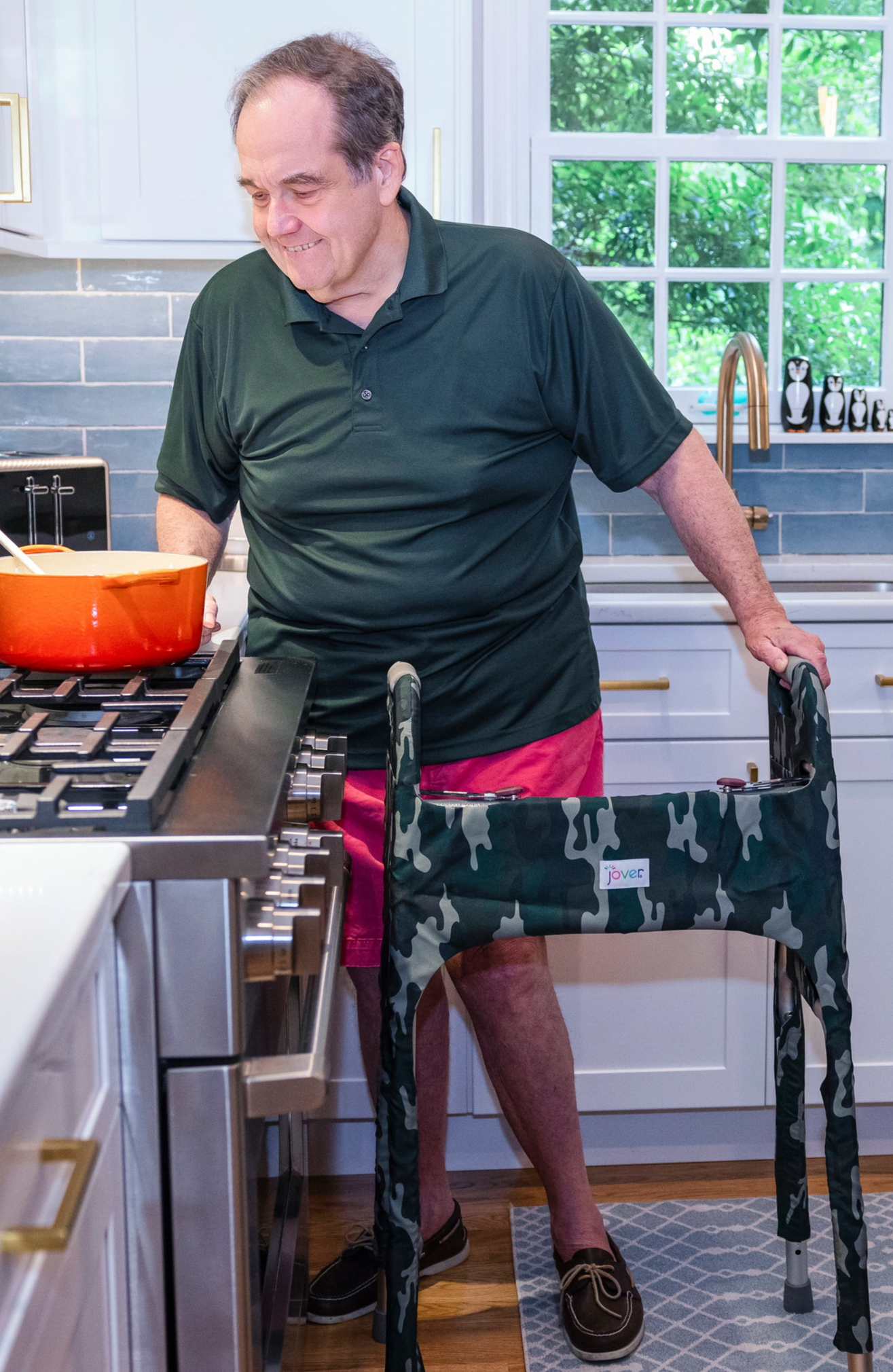 Man cooking in kitchen using a medical walker with a green and gray camouflage print walker cover on it. View highlights the tailored nature of the cover and the Jover logo on the front.