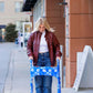 A young woman walking down a city sidewalk using a metal folding walker outfitted with the Jover Walker Cover in  a blue and white floral pattern. The ultimate walker accessory combines practicality, with four pockets, and personality with a variety of patterns and solids.