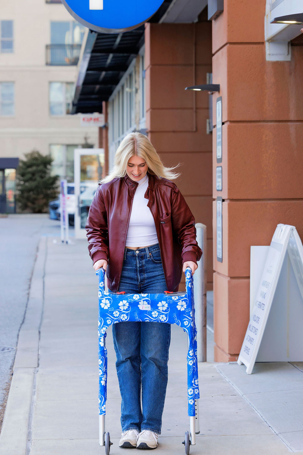 A young woman walking down a city sidewalk using a metal folding walker outfitted with the Jover Walker Cover in  a blue and white floral pattern. The ultimate walker accessory combines practicality, with four pockets, and personality with a variety of patterns and solids.