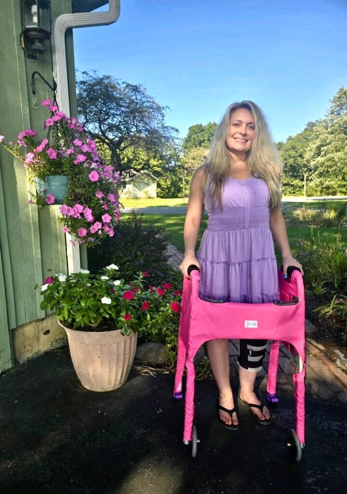 A woman smiles while standing on a pathway, using a standard folding walker with a bright pink Jover Walker Cover.