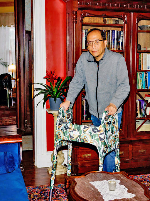 A man standing in front of a bookshelf in a historic home using a metal folding walker outfitted with a Jover Walker Cover in a camping inspired print called the Happy Camper.