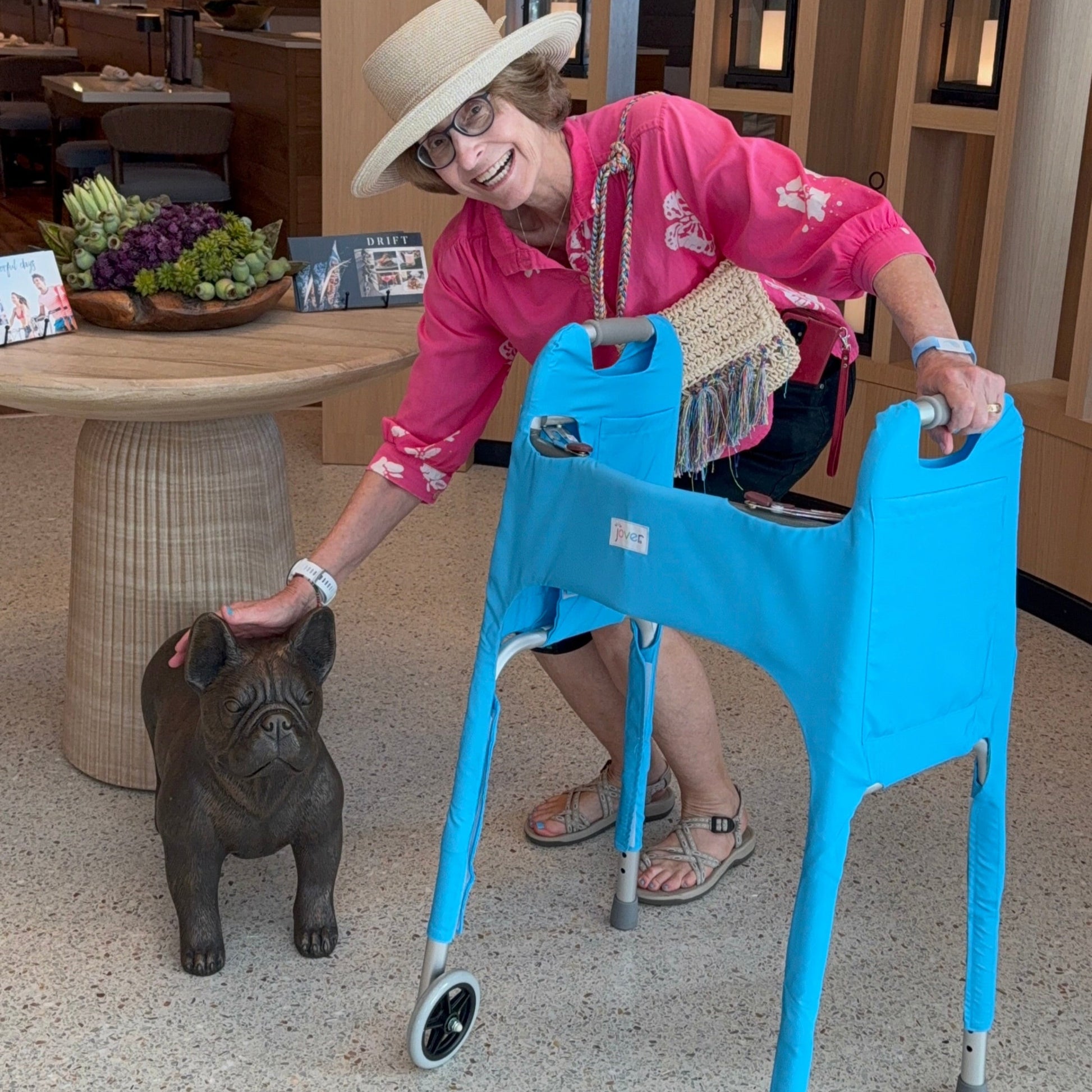 Woman in pink shirt and straw hat interacting with a small dog statue next to a standard walker clad in a sky blue Jover walker cover in an indoor setting.