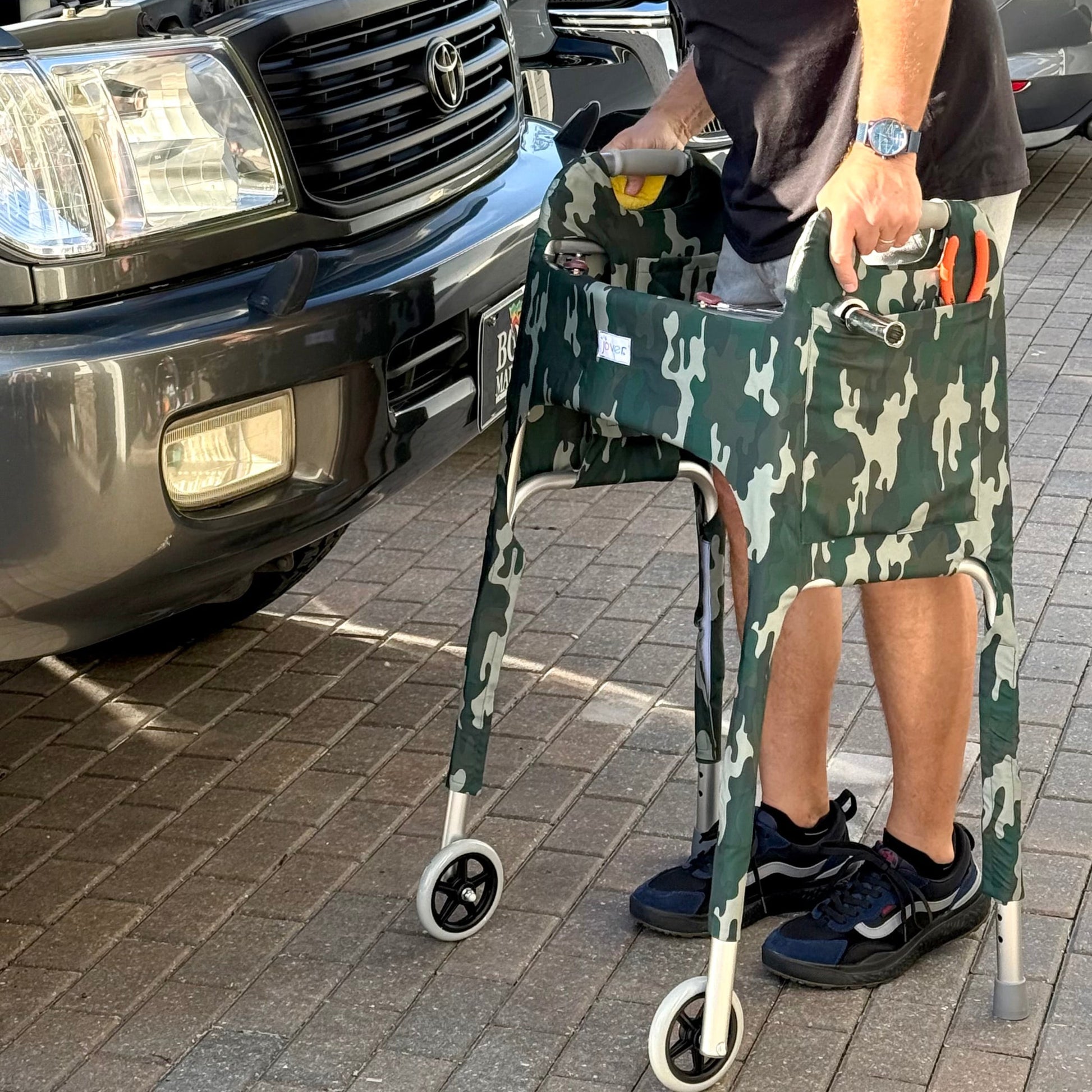 Man working on a car standing behind standard walker clad in Woodland Camo Jover Walker Cover with tools in the outside pocket.