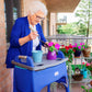A woman using a walker gardening in her patio. The walker sports a Jover Walker Cover in Deep Marine and in the side pocket she has an iPad. The walker also has a tray on it, which she is using to garden. The walker cover is compatible with other walker accessories.