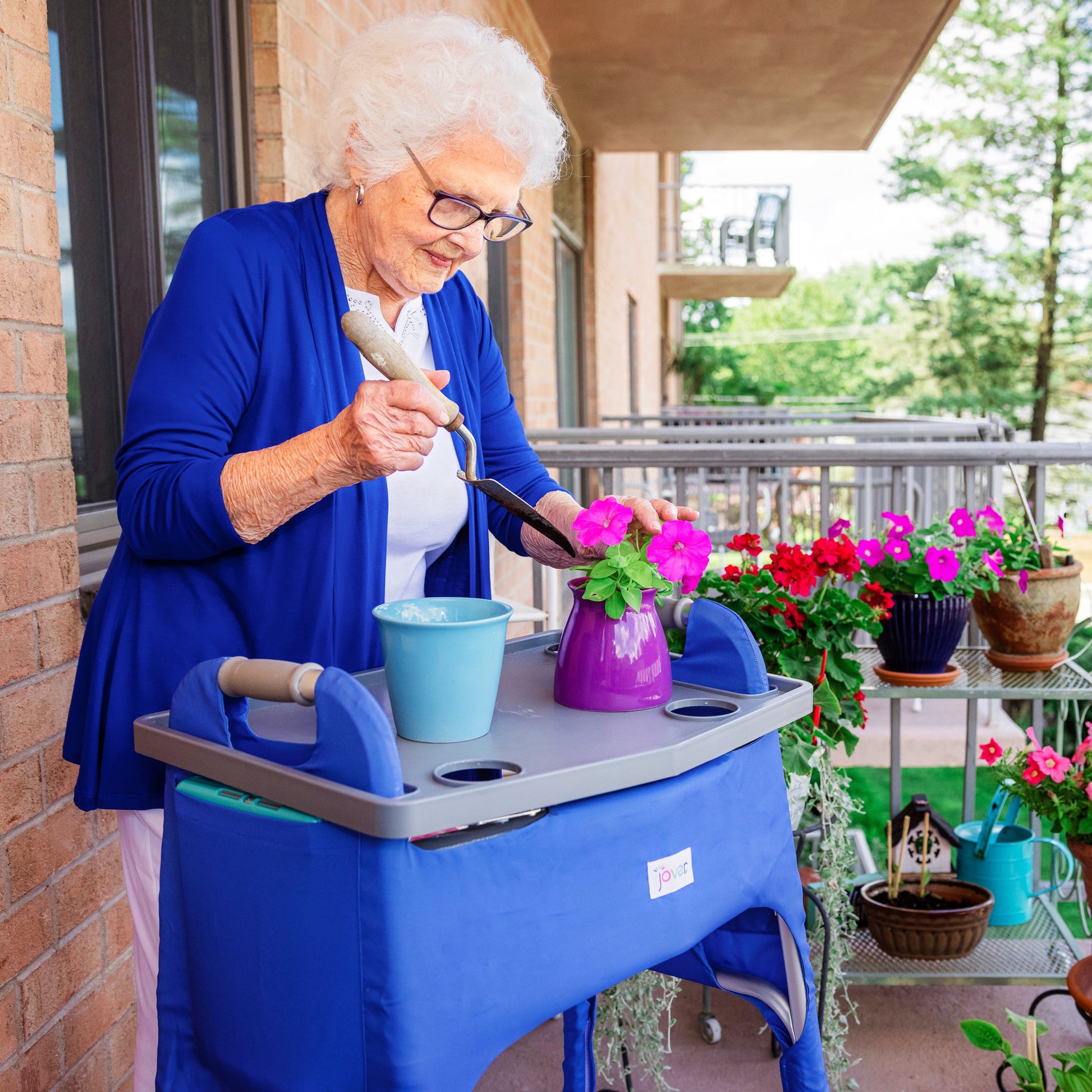 A woman using a walker gardening in her patio. The walker sports a Jover Walker Cover in Deep Marine and in the side pocket she has an iPad. The walker also has a tray on it, which she is using to garden. The walker cover is compatible with other walker accessories.