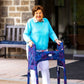 Woman in front of building smiling using a walker clad in the Jover Walker Cover in Fireworks--a dark blue backdrop covered in patriotic red, white, and blue bursts. With pockets and style, it's the perfect for anyone who loves the USA and a night sky fireworks show.