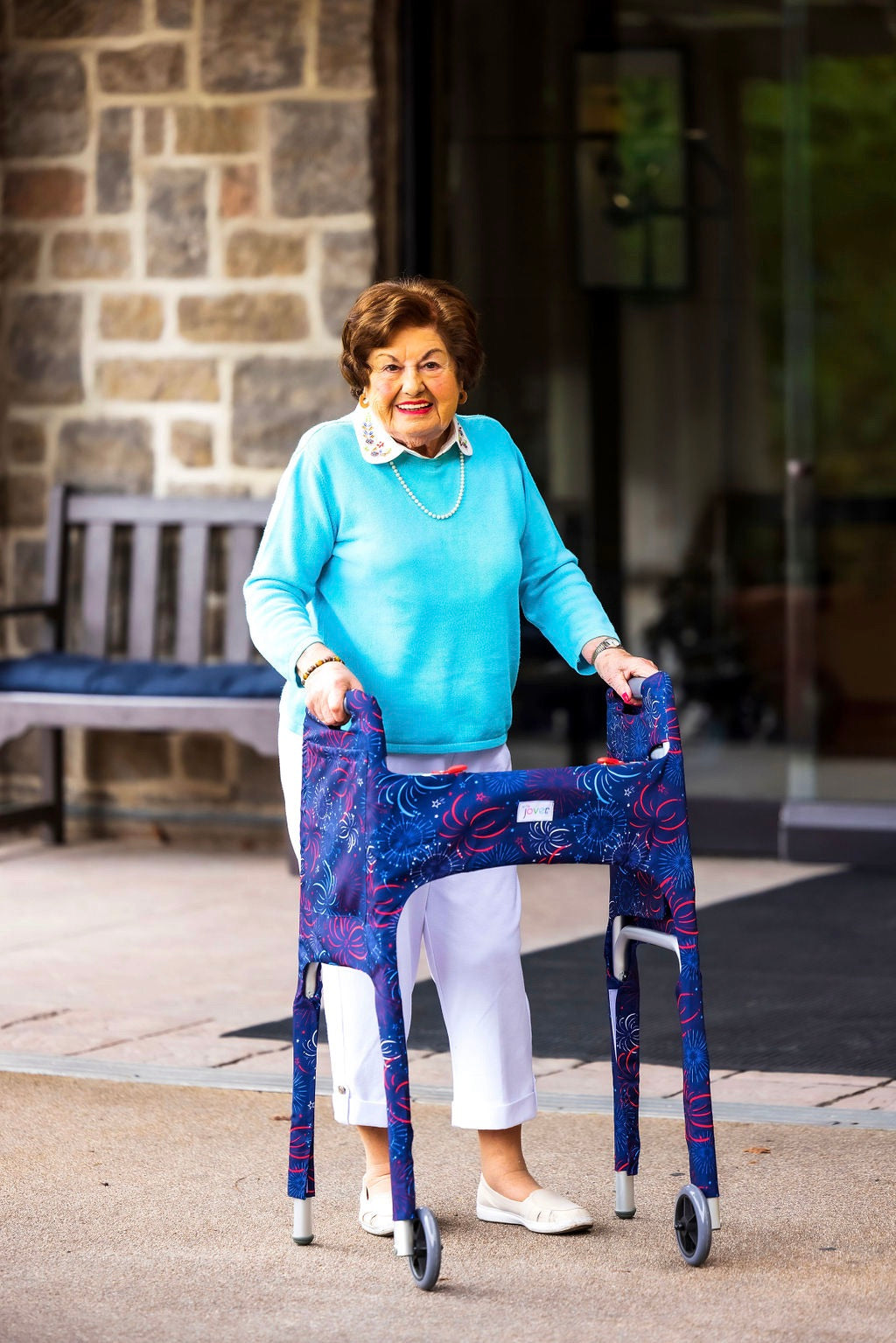 Woman in front of building smiling using a walker clad in the Jover Walker Cover in Fireworks--a dark blue backdrop covered in patriotic red, white, and blue bursts. With pockets and style, it's the perfect for anyone who loves the USA and a night sky fireworks show.