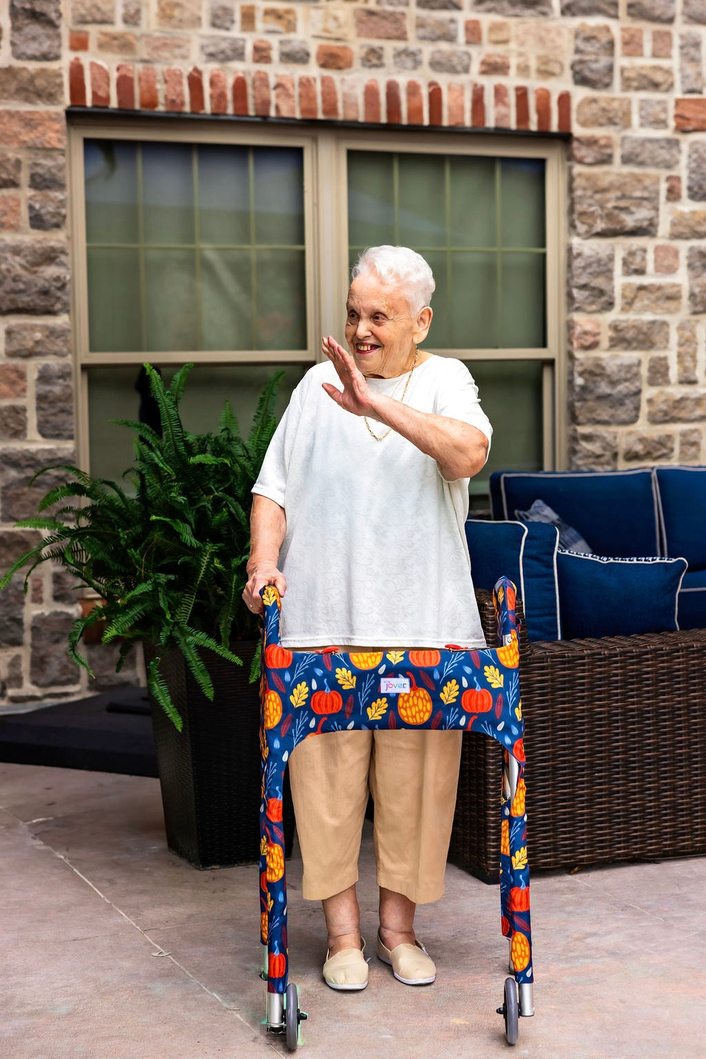 Woman on a patio waving and smiling using a walker clad in the Jover Walker Cover in Fall Festival--a dark blue backdrop covered in festive orange and yellow pumpkins and fall leaves. With pockets and style, it's the perfect fall accessory.