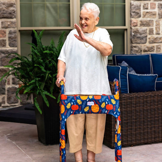 Woman on a patio waving and smiling using a walker clad in the Jover Walker Cover in Fall Festival--a dark blue backdrop covered in festive orange and yellow pumpkins and fall leaves. With pockets and style, it's the perfect fall accessory.