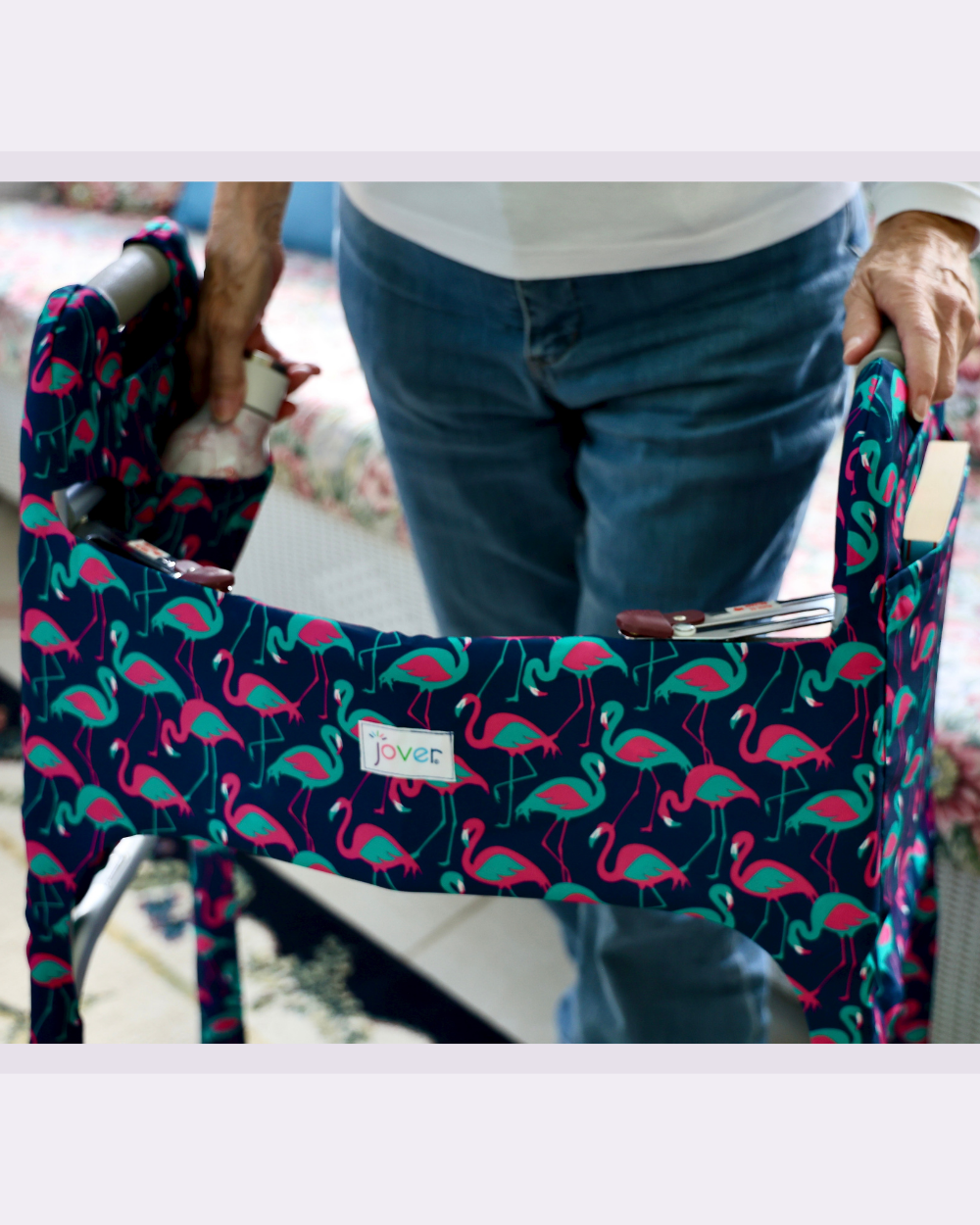 Front view of a woman standing behind a walker clad in a Jover Walker Cover in Flamingle print showing a book in an outside pocket and a water bottle in an inside pocket.